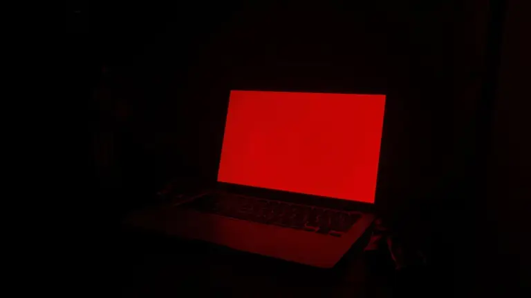 A laptop glowing red in a dark room beside leather gloves